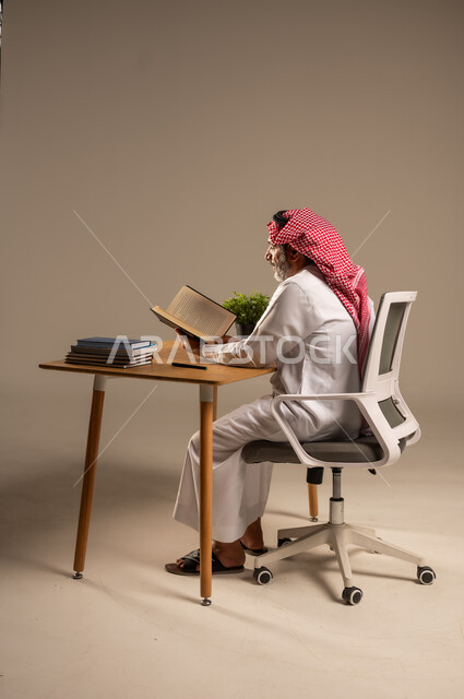 مهن ووظائف مكتبيه سعودية، استخدام التقنيات الحديثة والمتطورة في العمل، بورتريه لرجل كبير السن عربي خليجي سعودي يرتدي الثوب التقليدي والشماغ  يجلس خلف طاولة خشبية يعمل على جهاز الحاسوب, الجسم بالطول الكامل، خلفية بيج