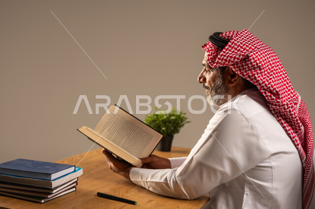  قضاء وقت الفراغ بالتعلم والثقافة، القراءة بتمعن وتركيز، بورتريه لرجل عربي سعودي خليجي كبير بالسن يرتدي الشماغ والثوب التقليدي يجلس على كرسي مريح يحمل بيده كتاب تثقيفي، خلفية بيج