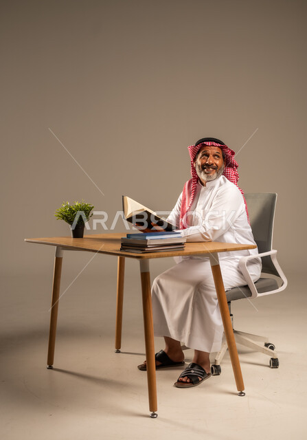  قضاء وقت الفراغ بالتعلم والثقافة، القراءة بتمعن وتركيز، بورتريه لرجل عربي سعودي خليجي كبير بالسن يرتدي الشماغ والثوب التقليدي يجلس على كرسي مريح يحمل بيده كتاب تثقيفي، خلفية بيج