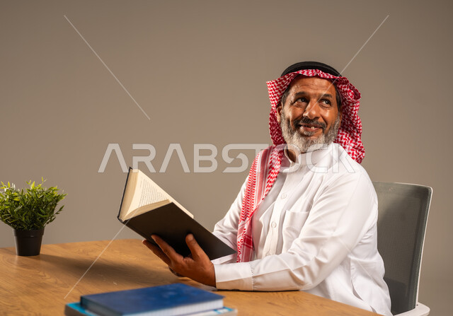  قضاء وقت الفراغ بالتعلم والثقافة، القراءة بتمعن وتركيز، بورتريه لرجل عربي سعودي خليجي كبير بالسن يرتدي الشماغ والثوب التقليدي يجلس على كرسي مريح يحمل بيده كتاب تثقيفي، خلفية بيج