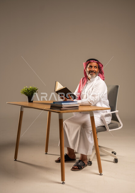  قضاء وقت الفراغ بالتعلم والثقافة، القراءة بتمعن وتركيز، بورتريه لرجل عربي سعودي خليجي كبير بالسن يرتدي الشماغ والثوب التقليدي يجلس على كرسي مريح يحمل بيده كتاب تثقيفي، خلفية بيج