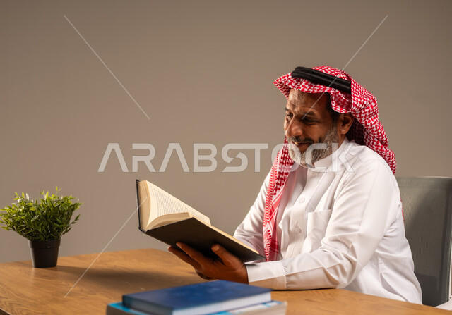  قضاء وقت الفراغ بالتعلم والثقافة، القراءة بتمعن وتركيز، بورتريه لرجل عربي سعودي خليجي كبير بالسن يرتدي الشماغ والثوب التقليدي يجلس على كرسي مريح يحمل بيده كتاب تثقيفي، خلفية بيج
