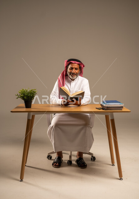  قضاء وقت الفراغ بالتعلم والثقافة، القراءة بتمعن وتركيز، بورتريه لرجل عربي سعودي خليجي كبير بالسن يرتدي الشماغ والثوب التقليدي يجلس على كرسي مريح يحمل بيده كتاب تثقيفي، خلفية بيج