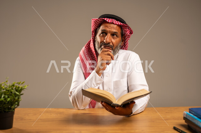  قضاء وقت الفراغ بالتعلم والثقافة، القراءة بتمعن وتركيز، بورتريه لرجل عربي سعودي خليجي كبير بالسن يرتدي الشماغ والثوب التقليدي يجلس على كرسي مريح يحمل بيده كتاب تثقيفي، خلفية بيج