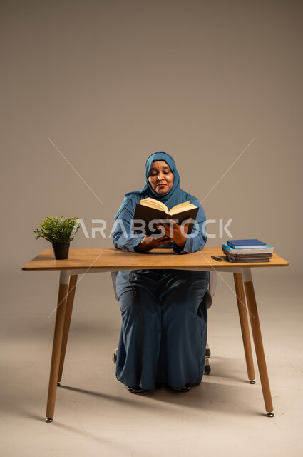 Reading carefully and with concentration, spending free time learning and culture, portrait of an elderly Saudi Arabian Gulf woman wearing a veil, wearing a navy blue abaya, sitting on a comfortable chair, holding an educational book in her hand, beige background