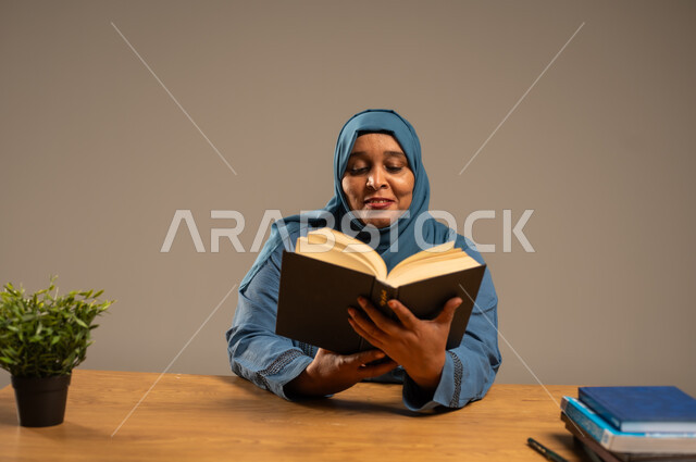 القراءة بتمعن وتركيز، قضاء وقت الفراغ بالتعلم والثقافة، بورتريه لإمراة  كبيرة بالسن عربية سعودية خليجية محجبة ترتدي العباءة الكحلية تجلس على كرسي مريح تحمل بيدها كتاب تثقيفي، خلفية بيج