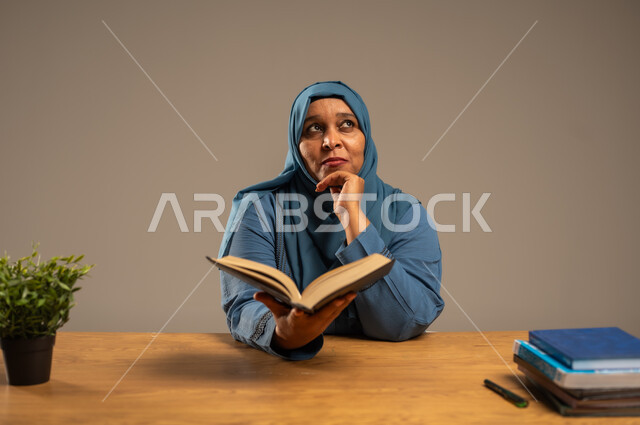 القراءة بتمعن وتركيز، قضاء وقت الفراغ بالتعلم والثقافة، بورتريه لإمراة  كبيرة بالسن عربية سعودية خليجية محجبة ترتدي العباءة الكحلية تجلس على كرسي مريح تحمل بيدها كتاب تثقيفي، خلفية بيج