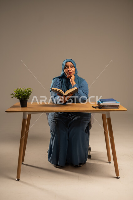 القراءة بتمعن وتركيز، قضاء وقت الفراغ بالتعلم والثقافة، بورتريه لإمراة  كبيرة بالسن عربية سعودية خليجية محجبة ترتدي العباءة الكحلية تجلس على كرسي مريح تحمل بيدها كتاب تثقيفي، خلفية بيج