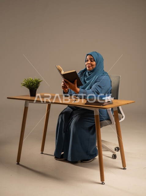 Reading carefully and with concentration, spending free time learning and culture, portrait of an elderly Saudi Arabian Gulf woman wearing a veil, wearing a navy blue abaya, sitting on a comfortable chair, holding an educational book in her hand, beige background