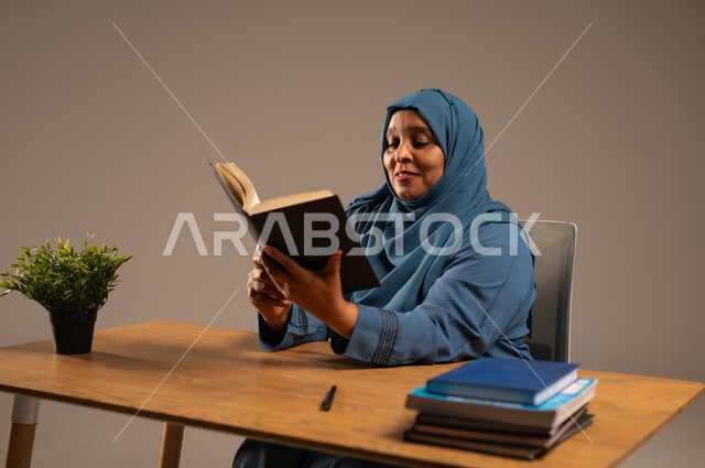 القراءة بتمعن وتركيز، قضاء وقت الفراغ بالتعلم والثقافة، بورتريه لإمراة  كبيرة بالسن عربية سعودية خليجية محجبة ترتدي العباءة الكحلية تجلس على كرسي مريح تحمل بيدها كتاب تثقيفي، خلفية بيج