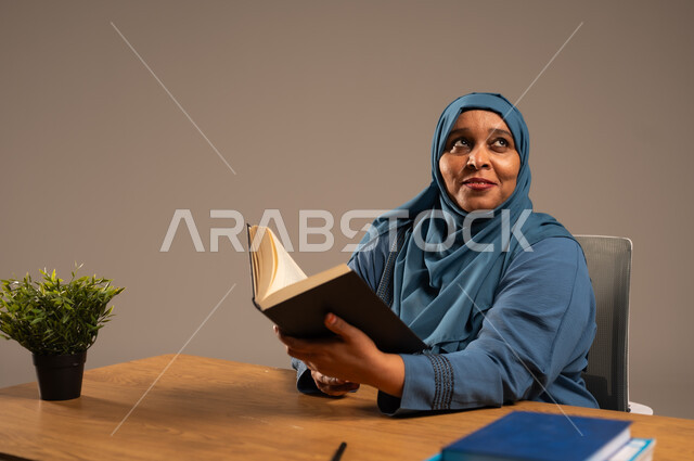 القراءة بتمعن وتركيز، قضاء وقت الفراغ بالتعلم والثقافة، بورتريه لإمراة  كبيرة بالسن عربية سعودية خليجية محجبة ترتدي العباءة الكحلية تجلس على كرسي مريح تحمل بيدها كتاب تثقيفي، خلفية بيج
