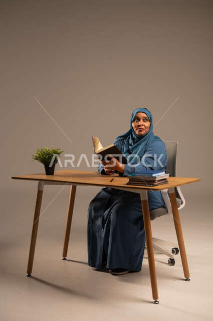 القراءة بتمعن وتركيز، قضاء وقت الفراغ بالتعلم والثقافة، بورتريه لإمراة  كبيرة بالسن عربية سعودية خليجية محجبة ترتدي العباءة الكحلية تجلس على كرسي مريح تحمل بيدها كتاب تثقيفي، خلفية بيج