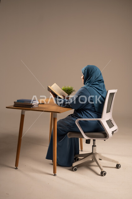 القراءة بتمعن وتركيز، قضاء وقت الفراغ بالتعلم والثقافة، بورتريه لإمراة  كبيرة بالسن عربية سعودية خليجية محجبة ترتدي العباءة الكحلية تجلس على كرسي مريح تحمل بيدها كتاب تثقيفي، خلفية بيج