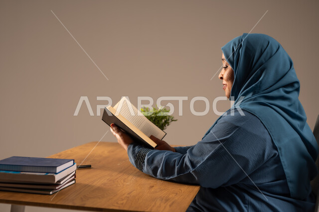 القراءة بتمعن وتركيز، قضاء وقت الفراغ بالتعلم والثقافة، بورتريه لإمراة  كبيرة بالسن عربية سعودية خليجية محجبة ترتدي العباءة الكحلية تجلس على كرسي مريح تحمل بيدها كتاب تثقيفي، خلفية بيج