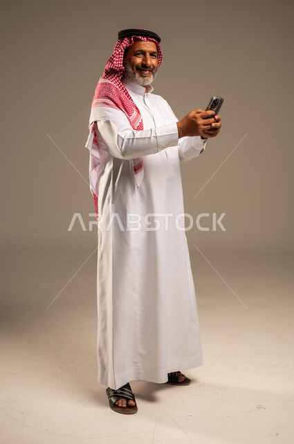 Development of technology and modern techniques, browsing social networking sites on the Internet, portrait of an elderly Saudi Arabian Gulf man wearing a traditional shemagh and thobe looking at the camera using a mobile phone, making video calls via mobile, full body image, beige background