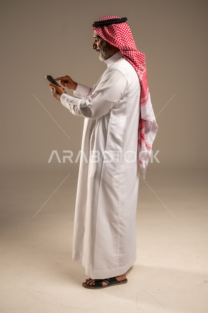 تطور التكنولوجيا والتقنيات الحديثة، تصفح مواقع التواصل الاجتماعي عبر الانترنت، بورتريه لرجل كبير السن عربي سعودي خليجي يرتدي الشماغ والثوب التقليدي يقوم باستخدام الهاتف المحمول، إجراء مكالمات فيديو عبر الجوال، صورة الجسم بالطول الكامل، خلفية بيج