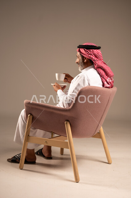 Mood enhancing caffeine, enjoying hot drinks, side portrait of elderly Saudi Arabian Gulf man wearing traditional keffiyeh and thobe sitting on chair holding cup of coffee, gestures of happiness and pleasure, beige background