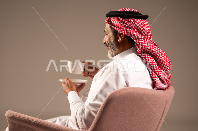 تعديل المزاج بشرب الكافيئين، الاستمتاع بتناول المشروبات الساخنة، بورتريه من الجانب لرجل مسن عربي خليجي سعودي يرتدي الشماغ والثوب التقليدي يجلس على كرسي ويحمل كوب من القهوة، ايماءات السعادة والسرور، خلفية بيج