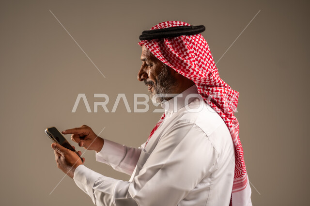 Development of technology and modern techniques, browsing social networking sites on the Internet, close-up portrait of an elderly Saudi Arabian Gulf man wearing a traditional shemagh and thobe using a mobile phone, making video calls via mobile, beige background