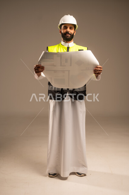 Concept of engineering and architectural construction, portrait of a Saudi Arabian Gulf engineer wearing traditional dress, special jacket and protective helmet holding a construction plan looking at the camera following up on the work, Saudi professions and jobs for young people, construction by the hands of the sons of the homeland, full body, beige background