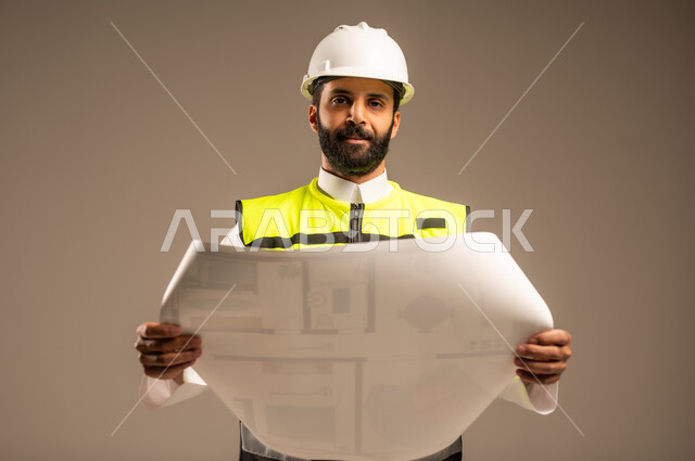 مفهوم الهندسة والانشاءات المعمارية، بورتريه مقرب لمهندس عربي خليجي سعودي يرتدي الثوب التقليدي والسترة الخاصة وخوذة الحماية يمسك مخطط الإعمار ينظر إلى الكاميرا يقوم بمتابعة الأعمال، مهن ووظائف سعودية للشباب، الإعمار بسواعد أبناء الوطن، خلفية بيج