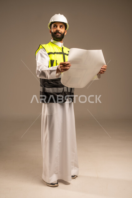Saudi jobs and professions for young people, construction by the hands of the sons of the homeland, the concept of engineering and architectural construction, a portrait of a Saudi Arabian Gulf engineer wearing the traditional dress, a special jacket and a protective helmet holding a construction plan, looking at something, following up on the work, the body in full length, beige background
