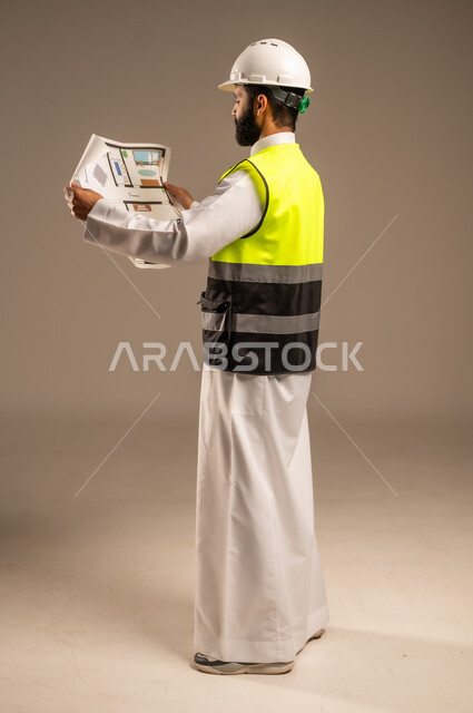 مهن ووظائف سعودية للشباب، الإعمار بسواعد أبناء الوطن، مفهوم الهندسة والانشاءات المعمارية، بورتريه من الجانب لمهندس عربي خليجي سعودي يرتدي الثوب التقليدي والسترة الخاصة وخوذة الحماية يمسك بيده مخطط الإعمار يقوم بمتابعة الأعمال، الجسم بالطول الكامل، خلفية بيج