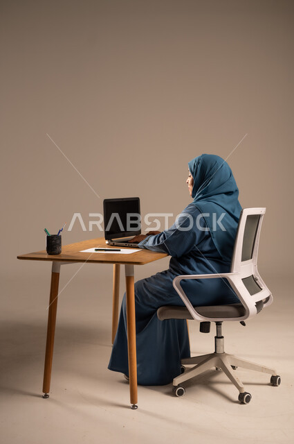 أداء المهام والاعمال المكتبيه داخل مقر الشركه، بورتريه لسيده  كبيرة بالسن عربيه خليجيه سعودية محجبة ترتدي العباءه الكحلية  تجلس خلف المكتب وتقوم بالعمل على جهاز الابتوب المحمول، دراسه المشاريع ومراجعه التقارير السنويه، جلسه عمل عن بعد، خلفيه بيج