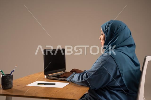أداء المهام والاعمال المكتبيه داخل مقر الشركه، بورتريه لسيده  كبيرة بالسن عربيه خليجيه سعودية محجبة ترتدي العباءه الكحلية  تجلس خلف المكتب وتقوم بالعمل على جهاز الابتوب المحمول، دراسه المشاريع ومراجعه التقارير السنويه، جلسه عمل عن بعد، خلفيه بيج