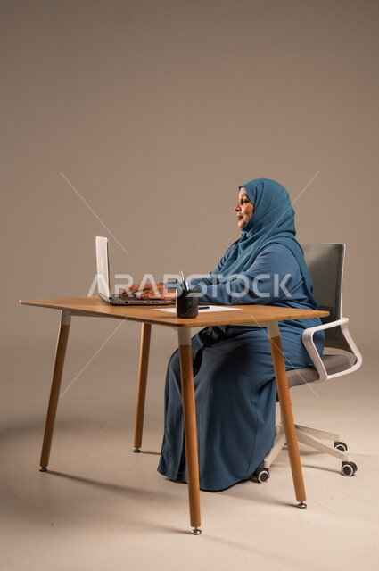 أداء المهام والاعمال المكتبيه داخل مقر الشركه، بورتريه لسيده  كبيرة بالسن عربيه خليجيه سعودية محجبة ترتدي العباءه الكحلية  تجلس خلف المكتب وتقوم بالعمل على جهاز الابتوب المحمول، دراسه المشاريع ومراجعه التقارير السنويه، جلسه عمل عن بعد، خلفيه بيج