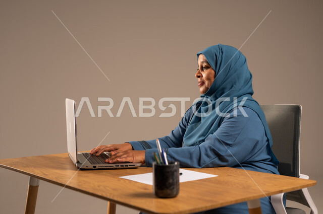 أداء المهام والاعمال المكتبيه داخل مقر الشركه، بورتريه لسيده  كبيرة بالسن عربيه خليجيه سعودية محجبة ترتدي العباءه الكحلية  تجلس خلف المكتب وتقوم بالعمل على جهاز الابتوب المحمول، دراسه المشاريع ومراجعه التقارير السنويه، جلسه عمل عن بعد، خلفيه بيج