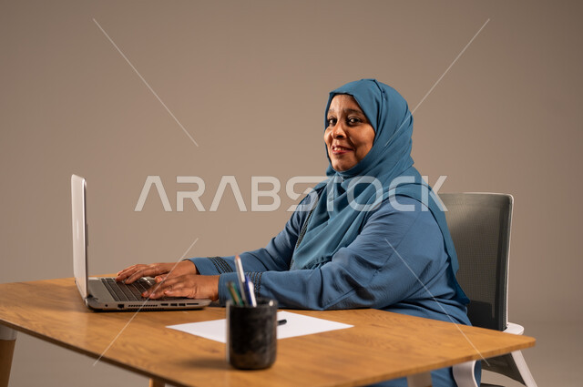 أداء المهام والاعمال المكتبيه داخل مقر الشركه، بورتريه لسيده  كبيرة بالسن عربيه خليجيه سعودية محجبة ترتدي العباءه الكحلية  تجلس خلف المكتب وتقوم بالعمل على جهاز الابتوب المحمول، دراسه المشاريع ومراجعه التقارير السنويه، جلسه عمل عن بعد، خلفيه بيج