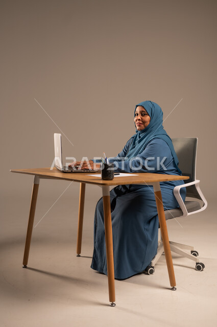 أداء المهام والاعمال المكتبيه داخل مقر الشركه، بورتريه لسيده  كبيرة بالسن عربيه خليجيه سعودية محجبة ترتدي العباءه الكحلية  تجلس خلف المكتب وتقوم بالعمل على جهاز الابتوب المحمول، دراسه المشاريع ومراجعه التقارير السنويه، جلسه عمل عن بعد، خلفيه بيج