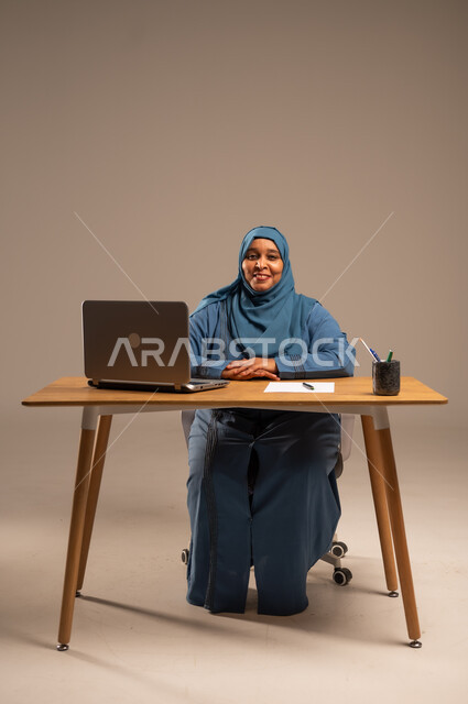أداء المهام والاعمال المكتبيه داخل مقر الشركه، بورتريه لسيده  كبيرة بالسن عربيه خليجيه سعودية محجبة ترتدي العباءه الكحلية  تجلس خلف المكتب وتقوم بالعمل على جهاز الابتوب المحمول، دراسه المشاريع ومراجعه التقارير السنويه، جلسه عمل عن بعد، خلفيه بيج