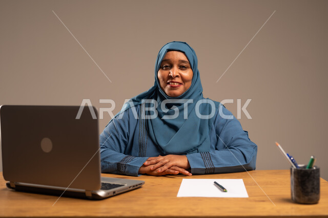 أداء المهام والاعمال المكتبيه داخل مقر الشركه، بورتريه لسيده  كبيرة بالسن عربيه خليجيه سعودية محجبة ترتدي العباءه الكحلية  تجلس خلف المكتب وتقوم بالعمل على جهاز الابتوب المحمول، دراسه المشاريع ومراجعه التقارير السنويه، جلسه عمل عن بعد، خلفيه بيج