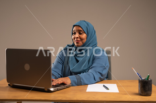 أداء المهام والاعمال المكتبيه داخل مقر الشركه، بورتريه لسيده  كبيرة بالسن عربيه خليجيه سعودية محجبة ترتدي العباءه الكحلية  تجلس خلف المكتب وتقوم بالعمل على جهاز الابتوب المحمول، دراسه المشاريع ومراجعه التقارير السنويه، جلسه عمل عن بعد، خلفيه بيج