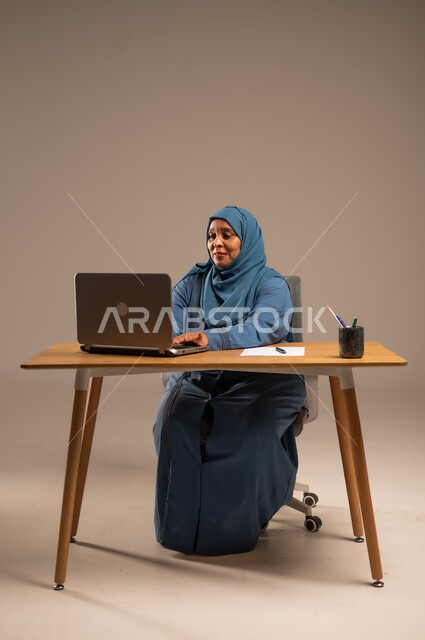 أداء المهام والاعمال المكتبيه داخل مقر الشركه، بورتريه لسيده  كبيرة بالسن عربيه خليجيه سعودية محجبة ترتدي العباءه الكحلية  تجلس خلف المكتب وتقوم بالعمل على جهاز الابتوب المحمول، دراسه المشاريع ومراجعه التقارير السنويه، جلسه عمل عن بعد، خلفيه بيج