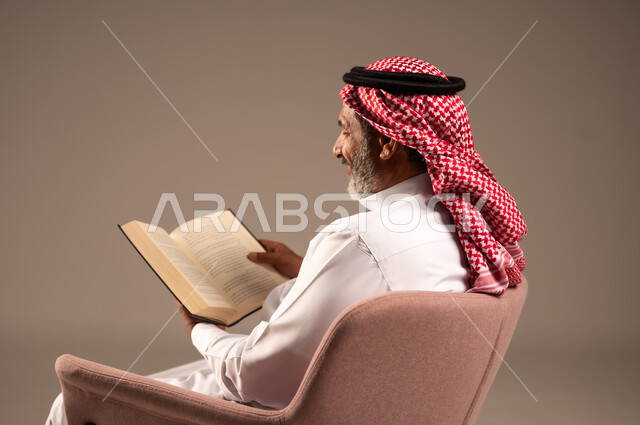 مفهوم محو الامية،  هواية حب المطالعة و الاستكشاف, قضاء وقت الفراغ بالتعلم والثقافة، بورتريه من الجانب لرجل كبير بالسن عربي خليجي سعودي  يرتدي الثوب التقليدي والشماغ يحمل بيده كتاب علمي تثقيفي، خلفية بيج