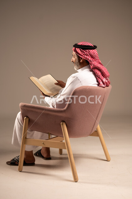 مفهوم محو الامية،  هواية حب المطالعة و الاستكشاف, قضاء وقت الفراغ بالتعلم والثقافة، بورتريه من الجانب لرجل كبير بالسن عربي خليجي سعودي  يرتدي الثوب التقليدي والشماغ يحمل بيده كتاب علمي تثقيفي، خلفية بيج