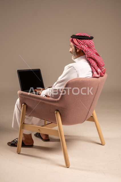 عقد إجتماع عمل عبر الإنترنت، تصفح برامج التواصل الاجتماعي, إستخدام الحاسوب المحمول لإنجاز المهام، بورتريه من الجانب لرجل عربي خليجي سعودي يرتدي الثوب التقليدي والشماغ يحمل جهاز اللابتوب بشاشة سوداء يجلس على كرسي مريح، تطور التكنولوجيا والأجهزة التقنية، خلفية بيج