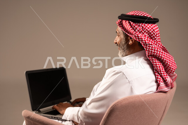 عقد إجتماع عمل عبر الإنترنت، تصفح برامج التواصل الاجتماعي, إستخدام الحاسوب المحمول لإنجاز المهام، بورتريه من الجانب لرجل عربي خليجي سعودي يرتدي الثوب التقليدي والشماغ يحمل جهاز اللابتوب بشاشة سوداء يجلس على كرسي مريح، تطور التكنولوجيا والأجهزة التقنية، خلفية بيج