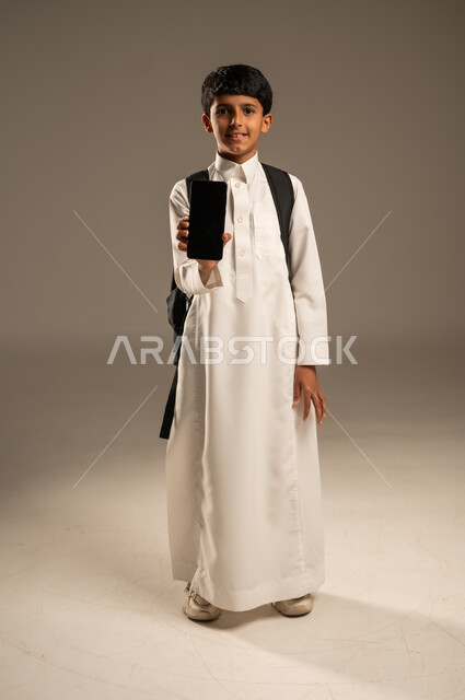 Using technological devices and techniques to display a blank black mobile screen, the concept of online education, a portrait of a Saudi Arabian Gulf boy wearing traditional dress and a school backpack holding a mobile phone in his hand and looking at the camera with gestures of pleasure, the development of learning methods and means, gray background