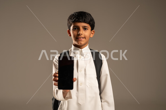Using technological devices and techniques to display a blank black mobile screen, the concept of online education, a portrait of a Saudi Arabian Gulf boy wearing traditional dress and a school backpack holding a mobile phone in his hand and looking at the camera with gestures of pleasure, the development of learning methods and means, gray background