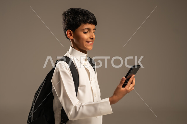 متابعة الحصص عبر الأونلاين, مفهوم التعليم عبر الانترنت, استخدام الأجهزة والتقنيات التكنولوجية, بورتريه من الجانب لولد عربي خليجي سعودي يرتدي الثوب التقليدي وحقيبة الظهر المدرسية يمسك الهاتف المحمول بيده بإيماءات التركيز، تطور وسائل وطرائق التعلم, خلفية رمادية
