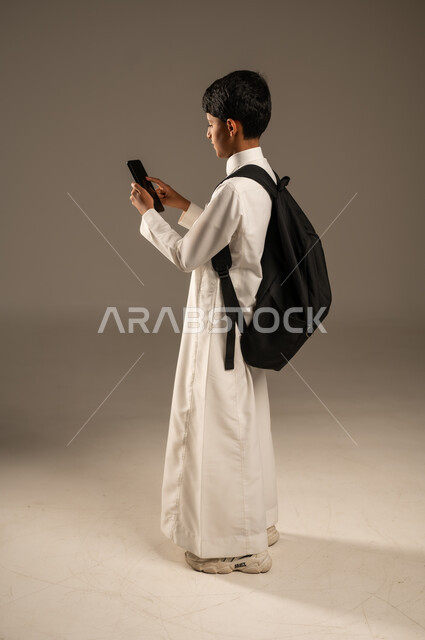 متابعة الحصص عبر الأونلاين, مفهوم التعليم عبر الانترنت, استخدام الأجهزة والتقنيات التكنولوجية, بورتريه من الجانب لولد عربي خليجي سعودي يرتدي الثوب التقليدي وحقيبة الظهر المدرسية يمسك الهاتف المحمول بيده بإيماءات التركيز، تطور وسائل وطرائق التعلم, خلفية رمادية