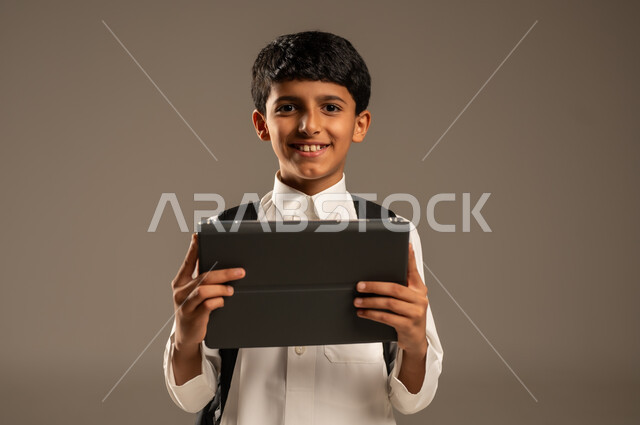 Online education concept, use of technological devices and techniques, following classes online, portrait of a Saudi Arabian Gulf boy wearing traditional dress and school backpack holding a tablet and looking at the camera with gestures of pleasure, development of learning methods and means, gray background