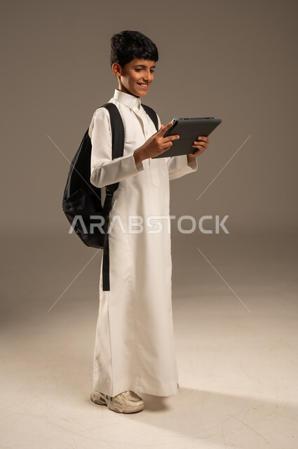 استخدام الأجهزة والتقنيات التكنولوجية الحديثة, مفهوم التعليم عبر الانترنت, متابعة الحصص عبر الأونلاين, بورتريه من الجانب لولد عربي خليجي سعودي يرتدي الثوب التقليدي يضع حقيبة الظهر المدرسية ويمسك بيده الجهاز اللوحي، تطور وسائل وطرائق التعلم, خلفية رمادية