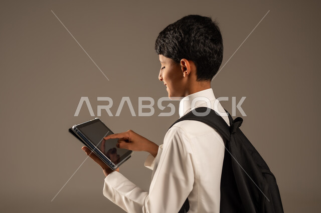 استخدام الأجهزة والتقنيات التكنولوجية الحديثة, مفهوم التعليم عبر الانترنت, متابعة الحصص عبر الأونلاين, بورتريه من الجانب لولد عربي خليجي سعودي يرتدي الثوب التقليدي يضع حقيبة الظهر المدرسية ويمسك بيده الجهاز اللوحي، تطور وسائل وطرائق التعلم, خلفية رمادية