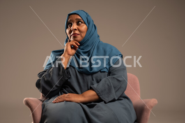  التأمل والتمعن والاندماج, بورتريه لإمرأة كبيرة بالسن عربية خليجية سعودية محجبة ترتدي العباءة الكحلية تجلس على الكرسي بايماءات وجه ويدين مختلفة، الثقة بالنفس والراحة النفسية، خلفية بيج