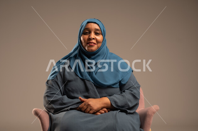  التأمل والتمعن والاندماج, بورتريه لإمرأة كبيرة بالسن عربية خليجية سعودية محجبة ترتدي العباءة الكحلية تجلس على الكرسي بايماءات وجه ويدين مختلفة، الثقة بالنفس والراحة النفسية، خلفية بيج